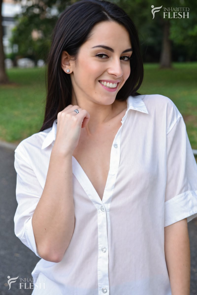Alt-text: "A woman in her 20s with a playful expression, wearing a white shirt, stands in a park on a sunny day.