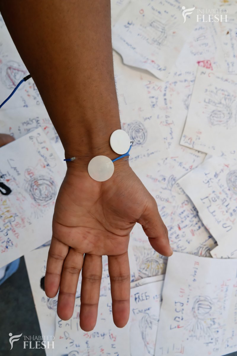 A man's hand with sensors connected, resting over discarded papers covered in equations and doodles, suggesting a scientific or experimental setting. The hand appears relaxed, with the palm facing up. The environment is indoors, possibly in a laboratory or workspace, with a focus on the hand and the surrounding papers.