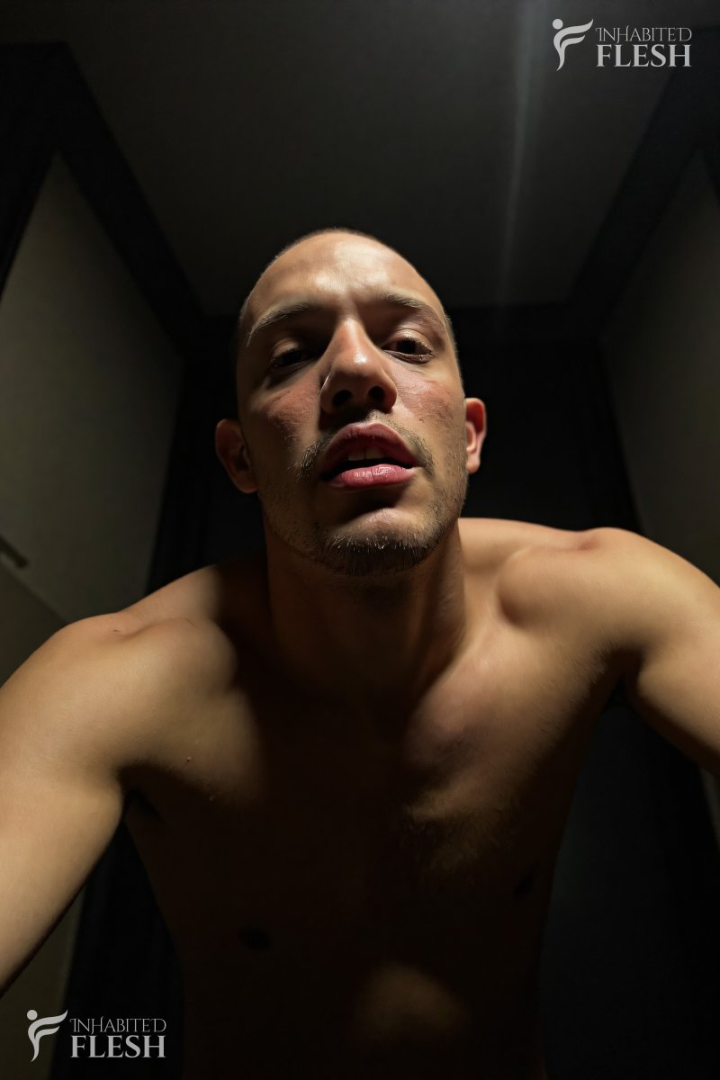 A shirtless man in his 20s, with a light complexion and short hair, looks directly at the viewer with a pleasure expression. He is standing in a dimly lit room, possibly a bedroom, with a neutral expression and no visible clothing. The setting appears to be indoors, likely at night.